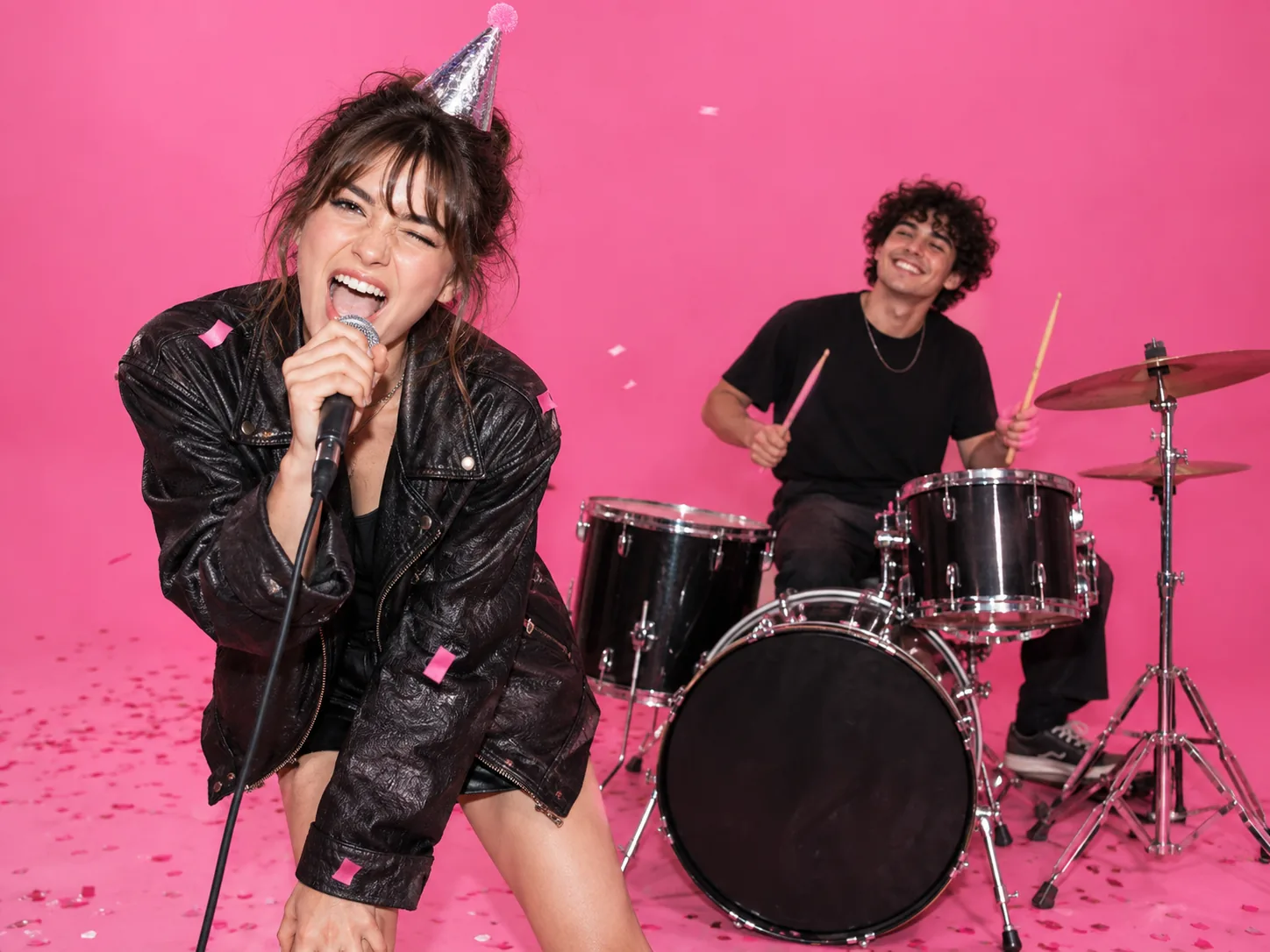 Birthday singer in a silver party hat and a friend on drums in a bright pink studio