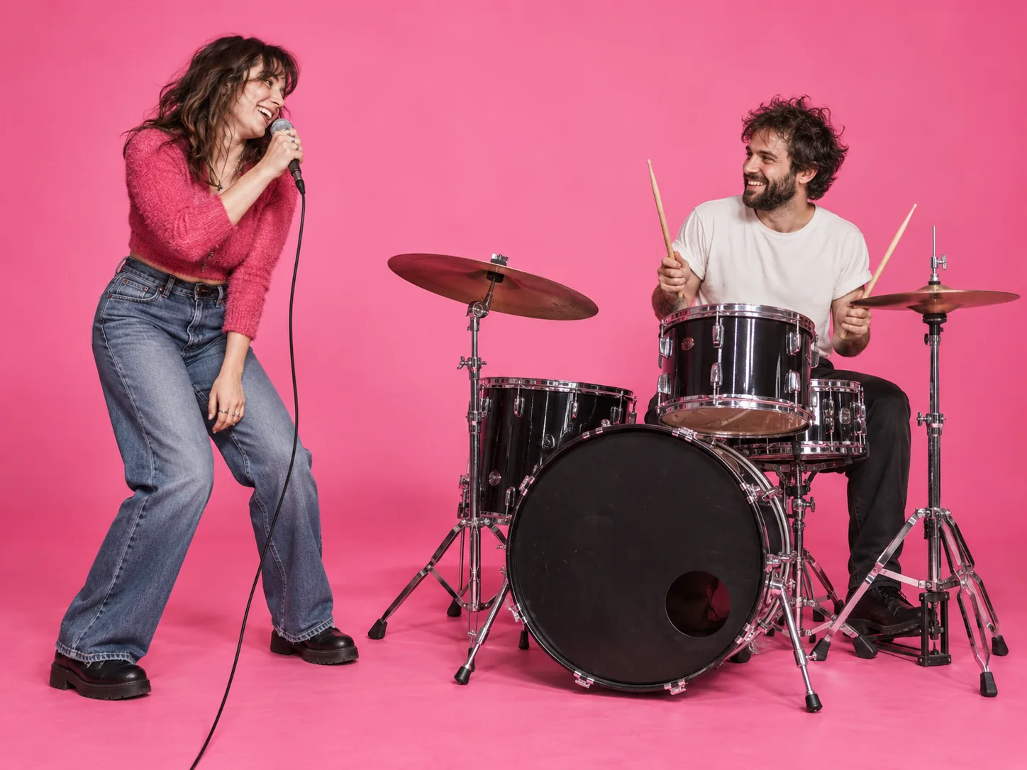 Two friends smiling at each other while one sings and the other drums in a pink studio